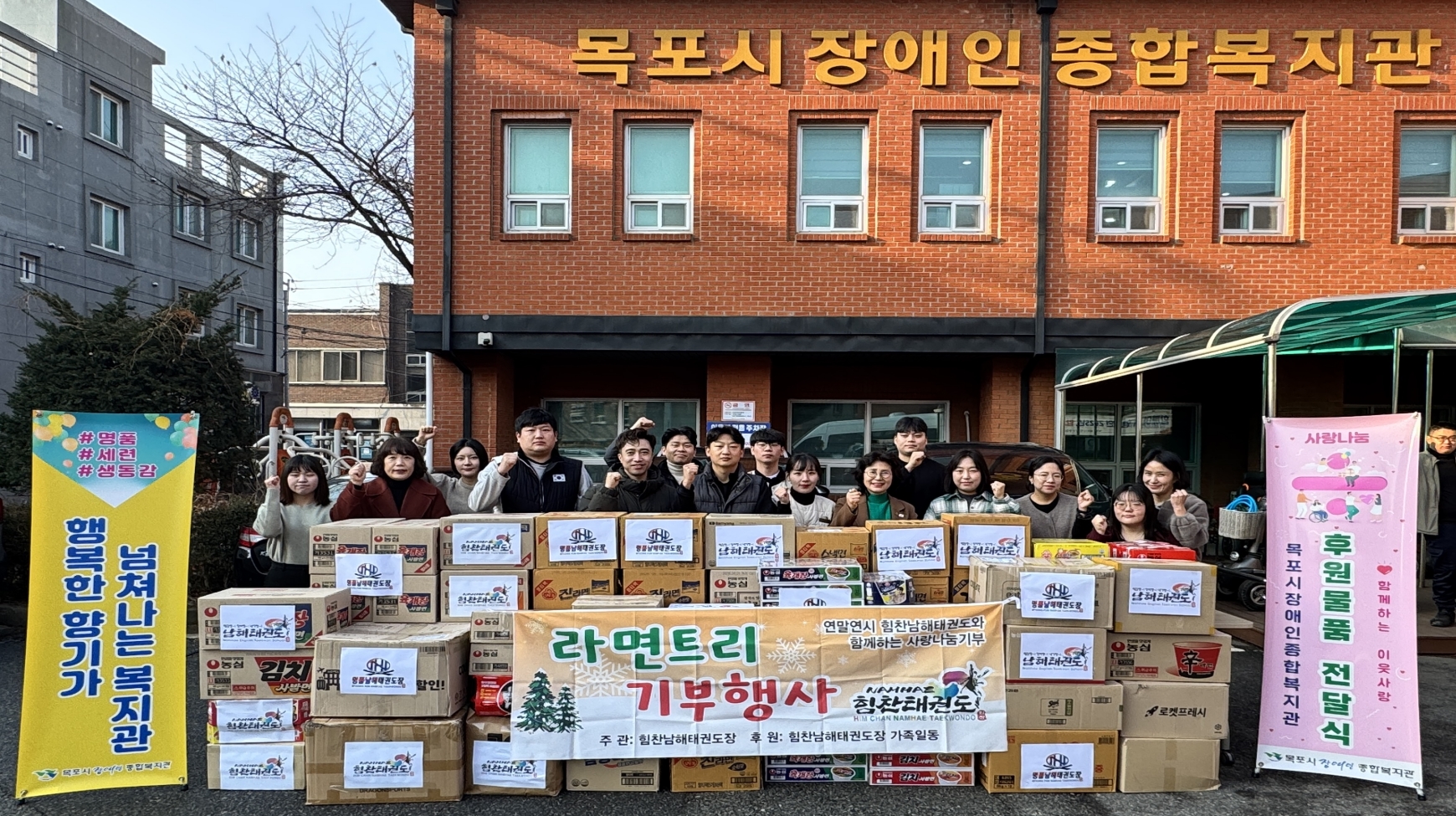 힘찬남해태권도장 관장과 수련생들이 목포시장애인종합복지관 앞마당에서 라면을 기부하고 단체 기념촬영을 하고 있다. 붉은 벽돌 건물인 복지관 외벽에는 '목포시 장애인 종합복지관'이라는 글자가 크게 붙어 있다. 앞쪽에는 기부된 라면 박스가 거대한 산처럼 쌓여 있고, 박스마다 태권도장 로고가 부착되어 있다. 라면 박스 앞에는 '라면트리 기부행사, 연말연시 힘찬남해태권도와 함께하는 사랑나눔기부, 주관: 힘찬남해태권도장'이라고 적힌 현수막이 걸려 있다. 관계자들은 모두 오른손 주먹을 쥐고 힘찬 파이팅 포즈를 취하고 있다.